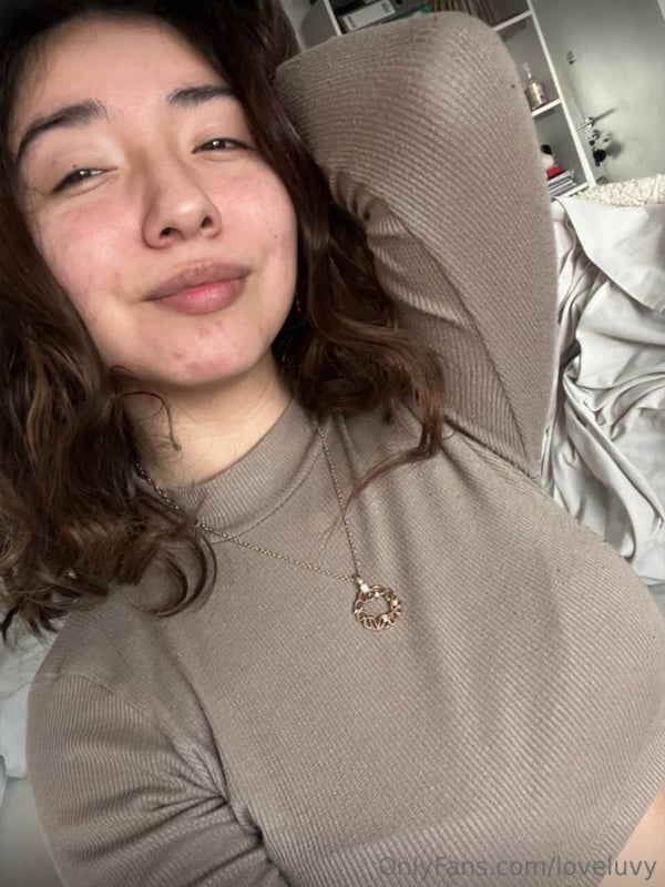 Young woman with wavy brown hair smiling playfully in a beige turtleneck top, arm raised against a room background.