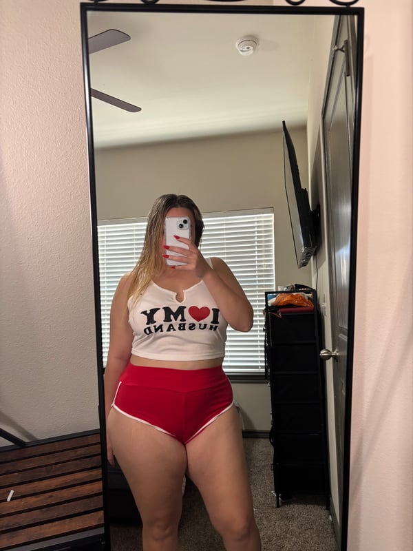 A woman standing in front of a mirror in a casual tank top and shorts, taking a selfie in a bedroom.