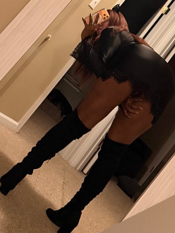 A woman posing playfully in a black lace top and thigh-high boots inside a room with beige walls and a door.