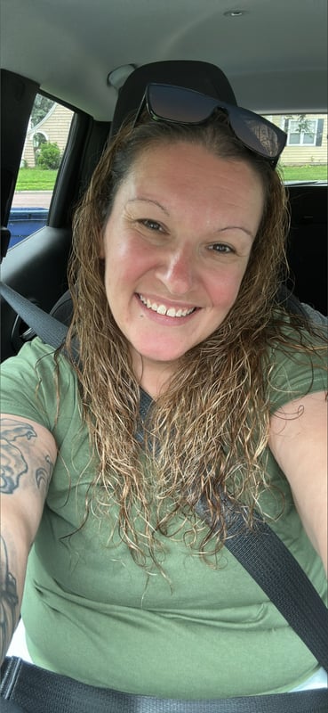 A smiling woman with curly hair and sunglasses perched on her head, sitting in a car wearing a green shirt, with visible tattoos on her arms.