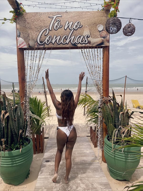 A woman in a white swimsuit posing with arms raised at a beach entrance decorated with tropical plants and a sign reading 'To To To Conconchasas' under a partly cloudy sky.