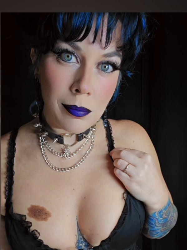 A woman with blue hair and bold makeup smiling at the camera, wearing layered necklaces, a black top, and plaid skirt against a dark background.