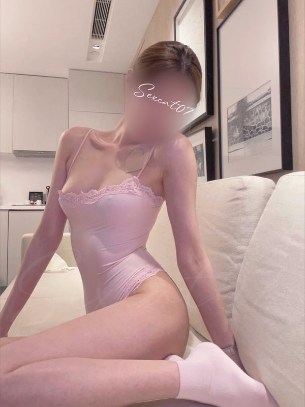 A woman in a pink lace bodysuit sitting on a couch in a modern living room with artwork on the walls.