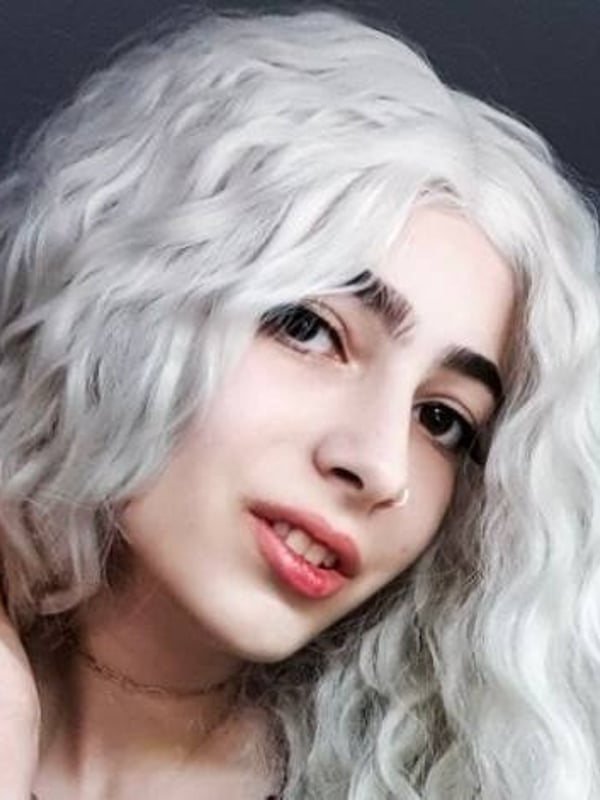 Close-up portrait of a person with long wavy white hair, subtle makeup, and a nose piercing, smiling softly.