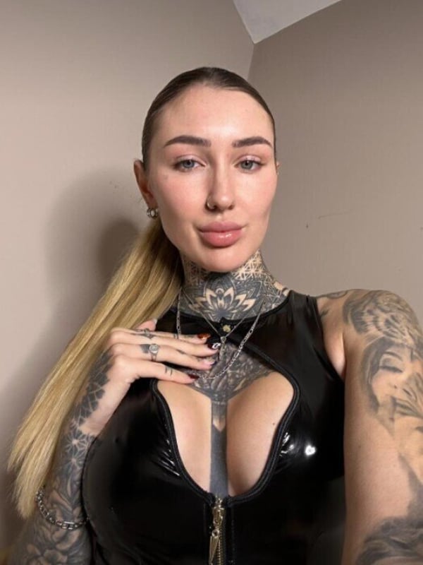 A woman with blonde hair in a ponytail, wearing a black shiny top, posing confidently indoors with tattoos visible.