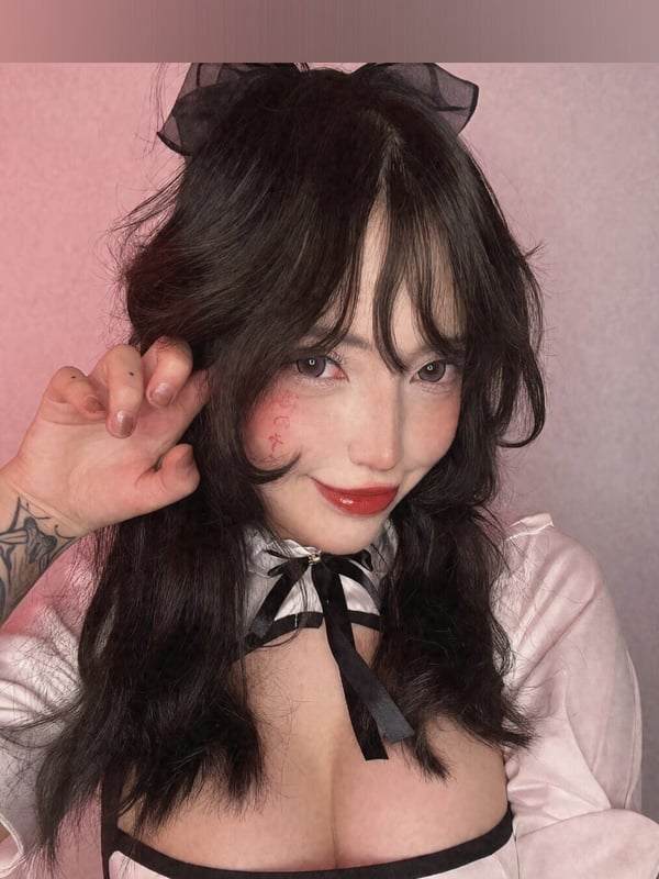 A person with long dark hair wearing a black bow and pink top against a soft pink backdrop.