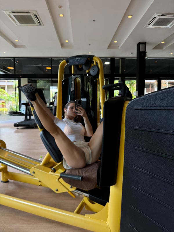 Young woman in athletic wear using a gym leg press machine, smiling at her phone in a modern fitness center.