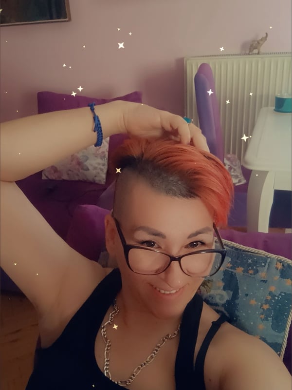 A woman with short dyed red hair and glasses smiling while sitting on purple cushions in a pink-toned room with artwork and lamps.