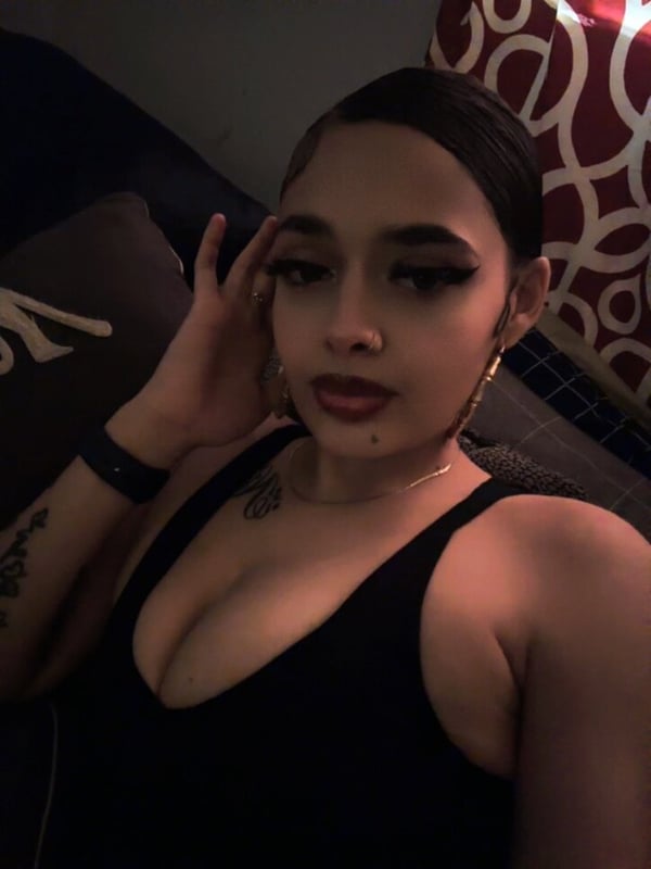 A woman with bold makeup sitting on a bed, wearing a black top and hoop earrings, with tattoos visible on her arm.