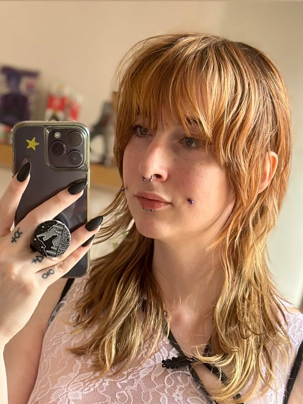 A person with shoulder-length light brown hair and facial piercings taking a selfie indoors while holding a phone.