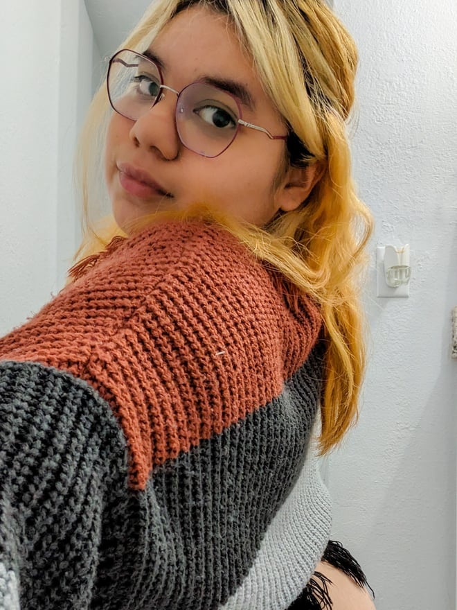 Young woman with blonde hair and glasses wearing a colorful striped sweater, posing in a bathroom with a towel rack in the background.