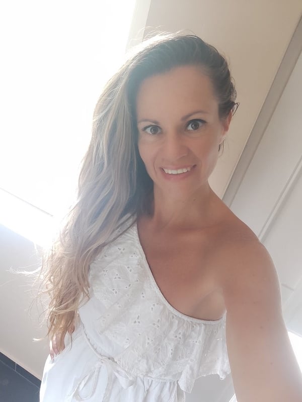 A smiling woman with long wavy hair wearing a white off-shoulder top, standing in a brightly lit room.