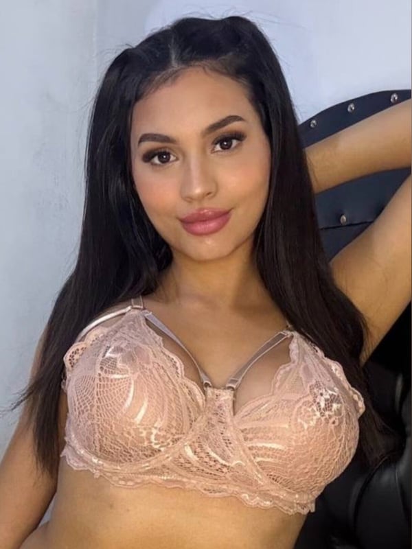 Young woman with long dark hair in pigtails, smiling at the camera while wearing a lace bra and posing indoors.