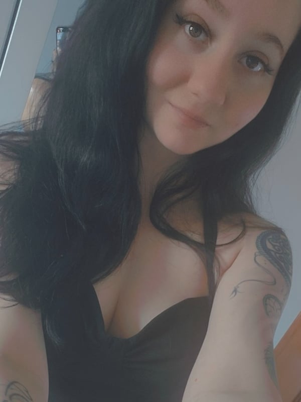 A young woman with long dark hair and tattoos smiles at the camera in a casual indoor setting.