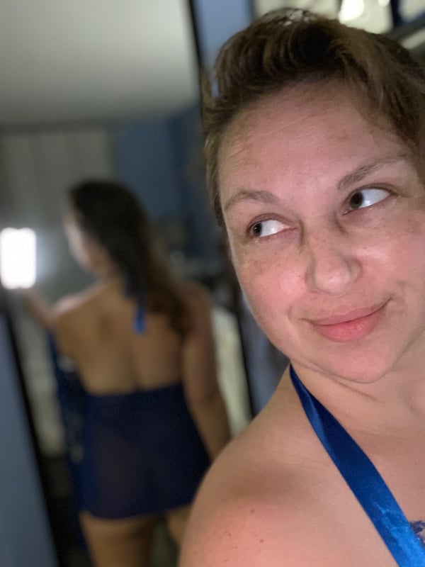 A woman with brunette hair smirking playfully at the mirror while wearing a sheer blue halter top and bottoms, standing in a bathroom.