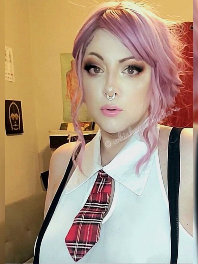 Woman with pink wavy hair, wearing a white collared top with suspenders and a plaid tie, posing indoors with a septum piercing.