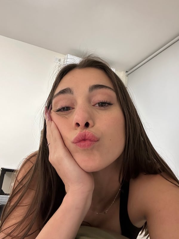 A close-up selfie of a person with long brown hair making a kissing face while resting their cheek on their hand.