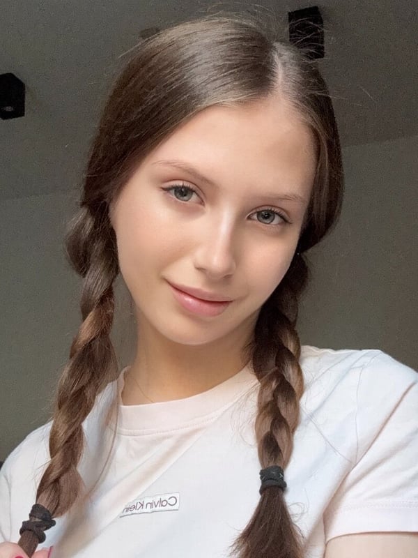 Close-up portrait of a young woman with brown hair in pigtails, wearing a light-colored top, smiling gently indoors.