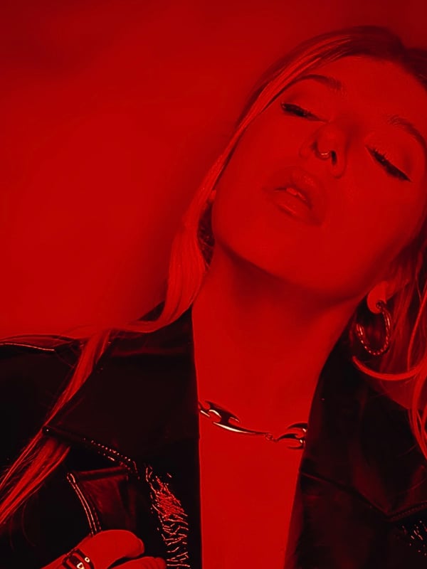 Close-up portrait of a woman with long blonde hair, wearing hoop earrings and a chain necklace, posing with eyes closed against a red background.