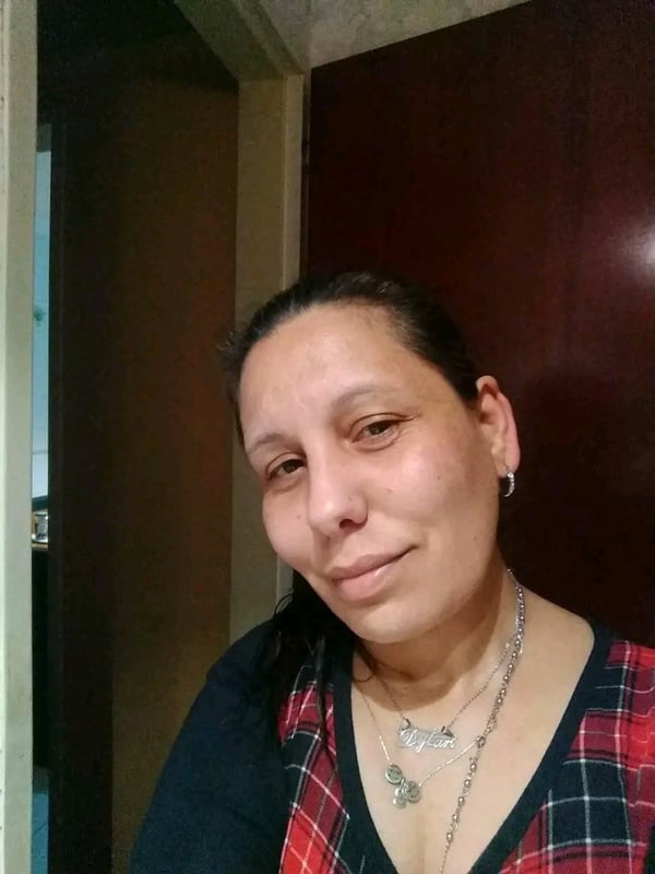 A woman with dark hair wearing a plaid shirt stands in a doorway with wooden doors and walls in the background.