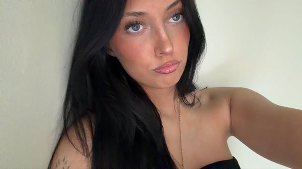 Young woman with long dark hair taking a mirror selfie in a bedroom, wearing a white top and gray shorts while sitting on a bed.