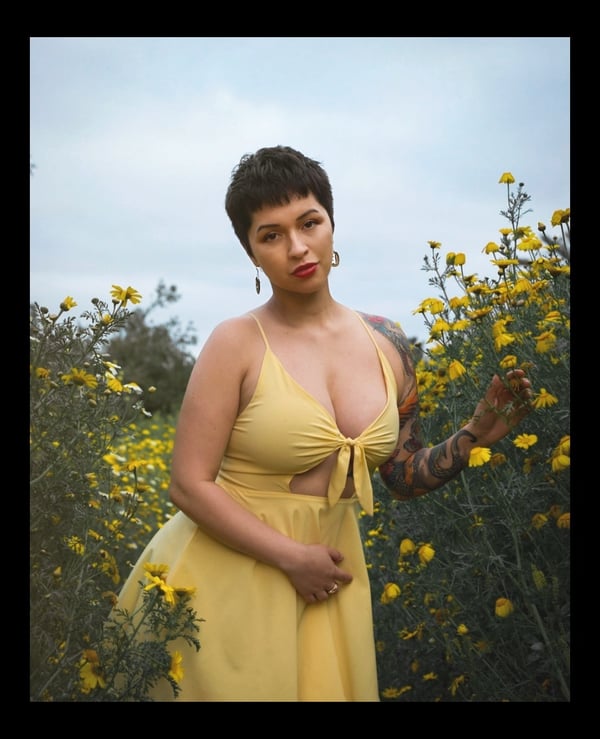 Woman with short dark hair wearing a yellow sleeveless dress, standing amidst yellow wildflowers under a cloudy sky.