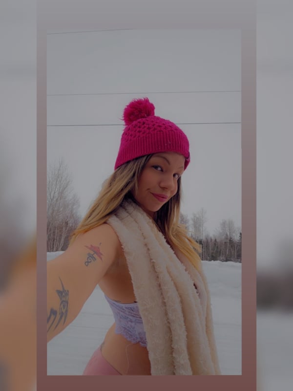 A smiling person outdoors in snowy weather, wearing a pink beanie, white scarf, and light top, with blonde hair and a tattoo on the arm.