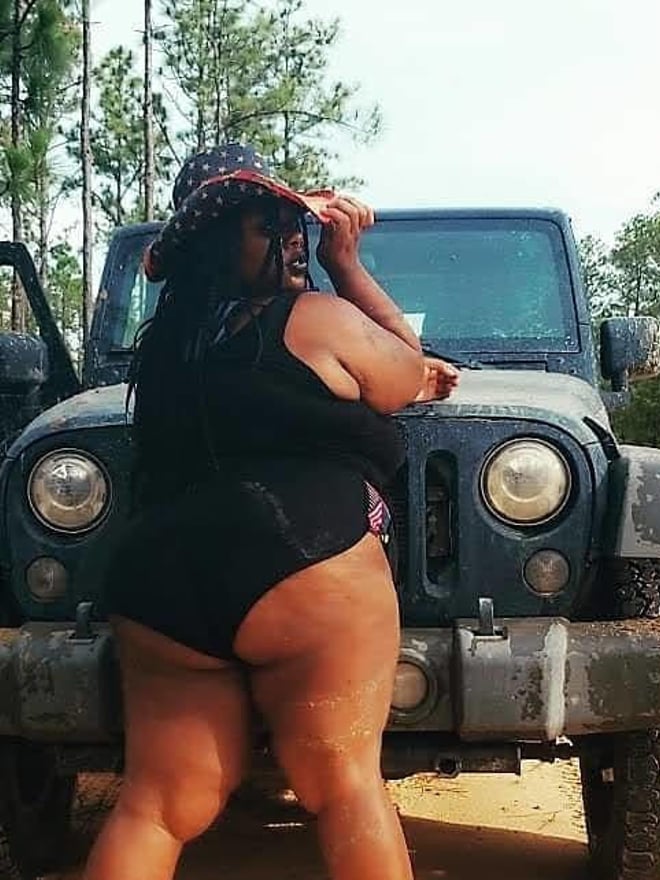 A woman in a black swimsuit posing next to a Jeep in a wooded outdoor area with pine trees, wearing a wide-brimmed hat.