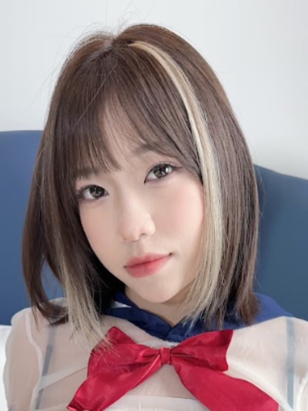 A young woman with shoulder-length hair featuring highlights, wearing a sheer top with a red bow.