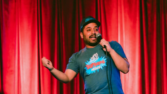 A man performing stand-up comedy on stage, holding a microphone and wearing a cap and graphic t-shirt against a red curtain backdrop.