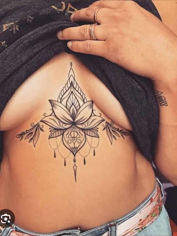 A woman posing in a loose gray top, revealing intricate floral tattoos on her chest and abdomen, with hands gently placed on her clothing.