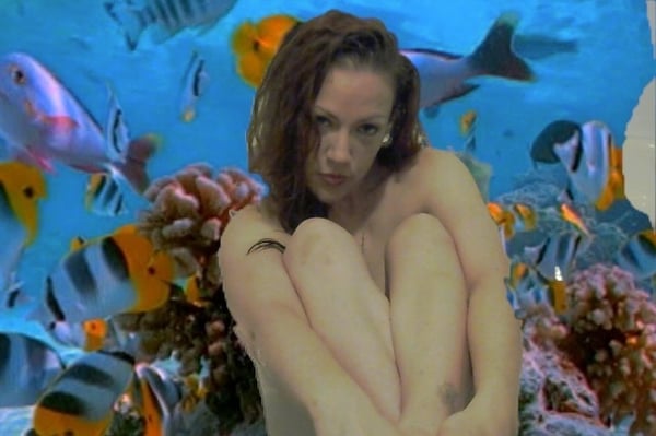 A woman with wavy brown hair sits with knees drawn up in front of a vibrant aquarium filled with colorful tropical fish and coral.