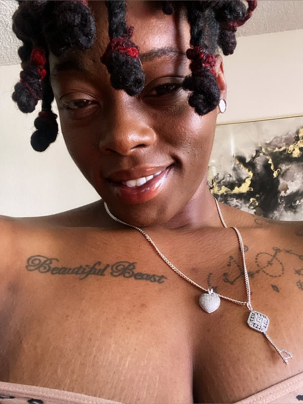 Close-up portrait of a smiling woman with braided hair and a necklace, showing tattoos on her chest.
