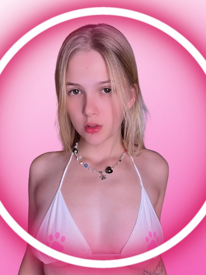 Close-up portrait of a young blonde woman with piercings and a necklace, smiling softly while wearing a white bikini top, framed in a glowing pink circle.