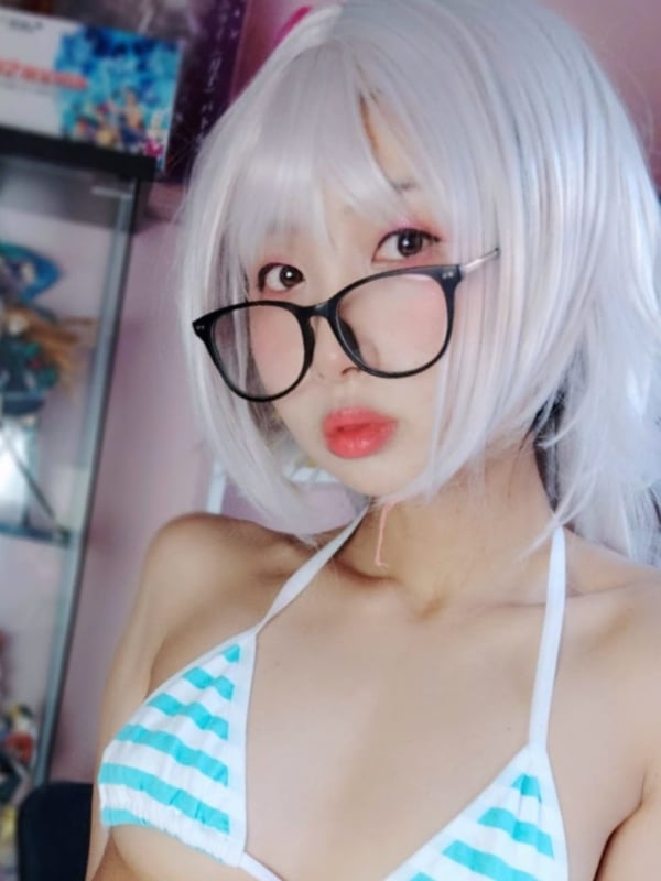 Young woman with white bob wig and glasses wearing a striped bikini top, posing playfully in a room with anime decor.