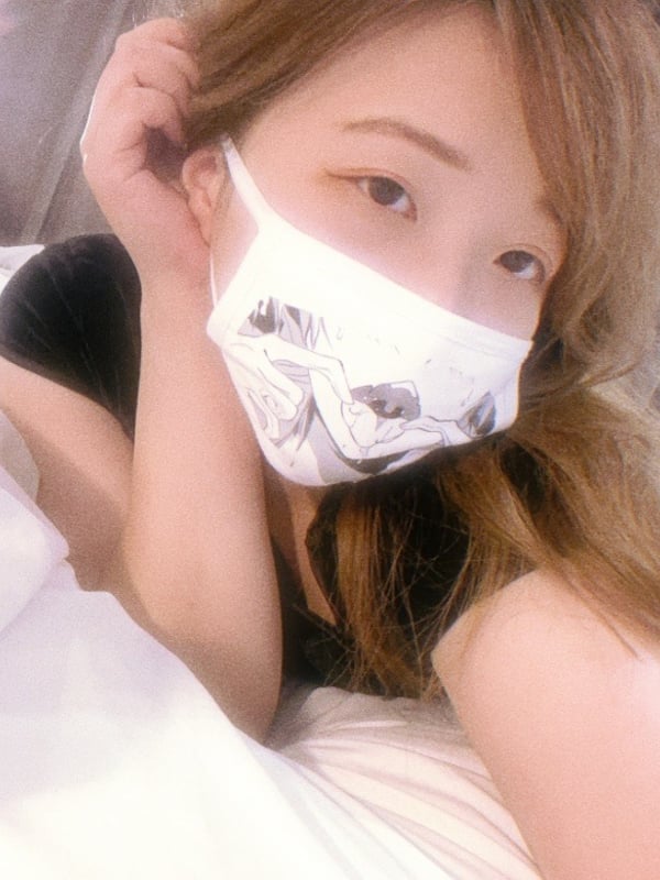 Young woman with long brown hair wearing a white mask with a black design, posing with hand near her face in a dimly lit setting.