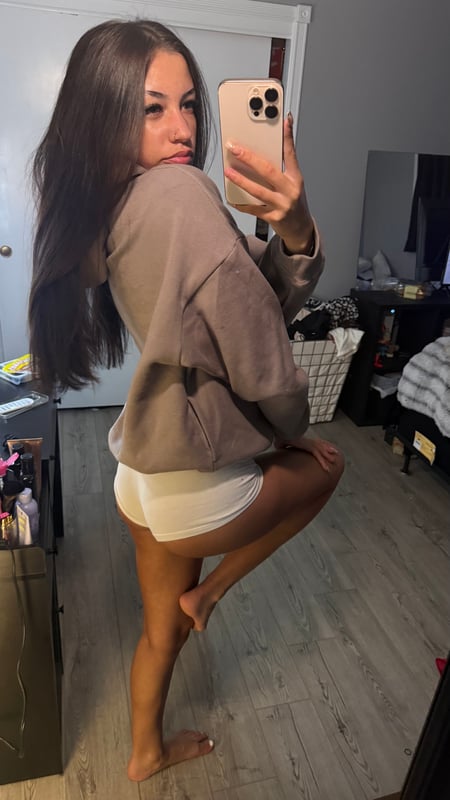 Young woman with long dark hair taking a mirror selfie in a beige tube top and jewelry, posing with hands near her face in a dimly lit room.
