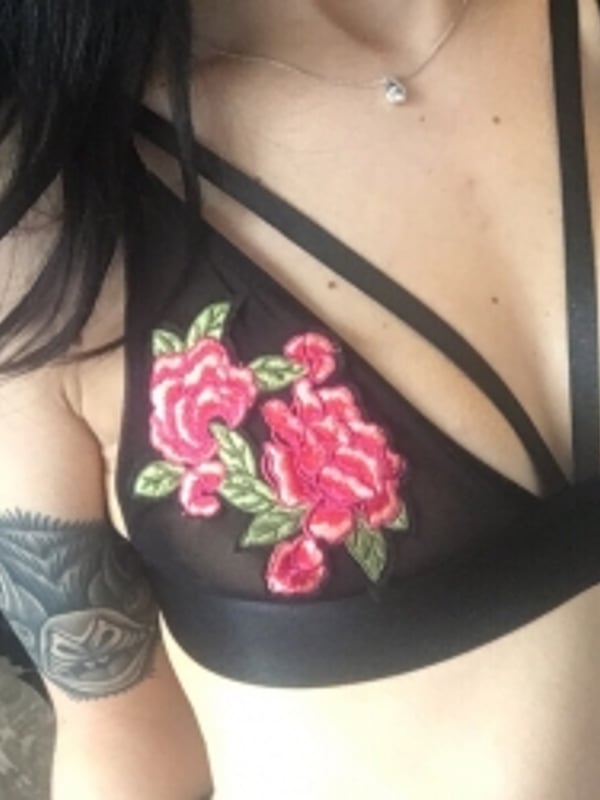 A woman posing in a black lace top with floral embroidery, wearing a delicate necklace.