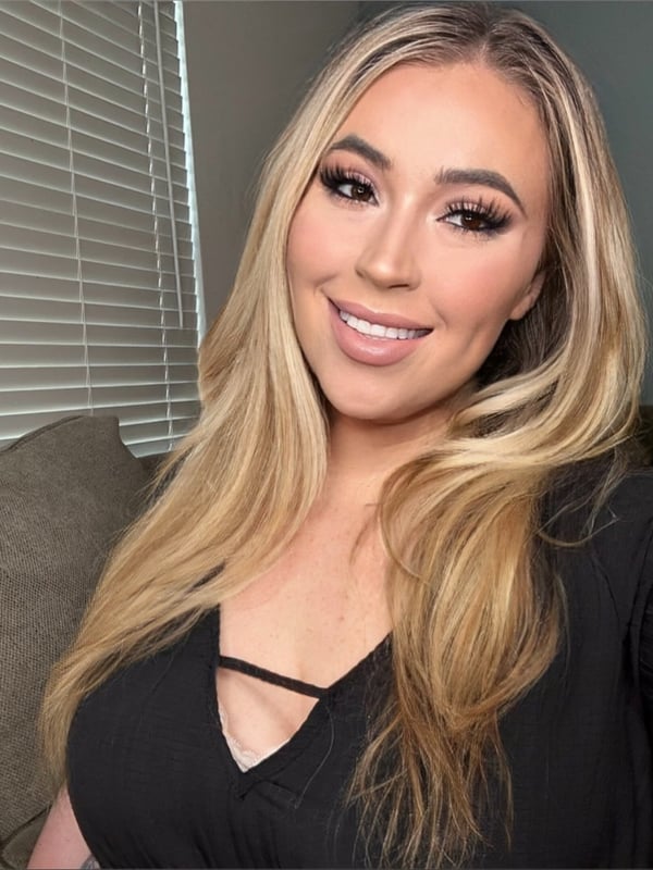 A smiling woman with long blonde hair taking a selfie in a black top inside a cozy room with beige furniture.