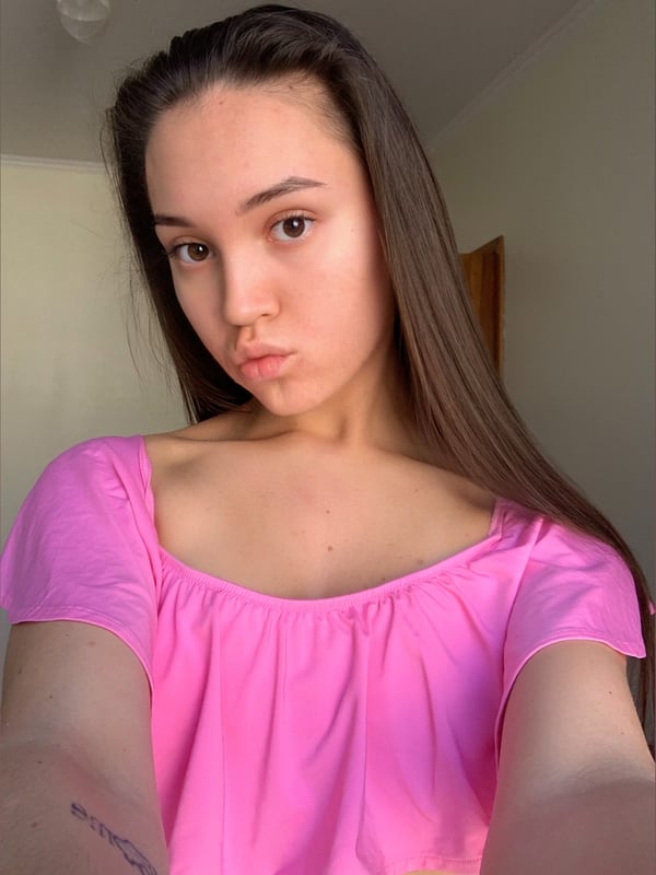 Close-up selfie of a young woman with long dark hair in a ponytail, wearing a pink off-shoulder top, pouting slightly in a brightly lit room.