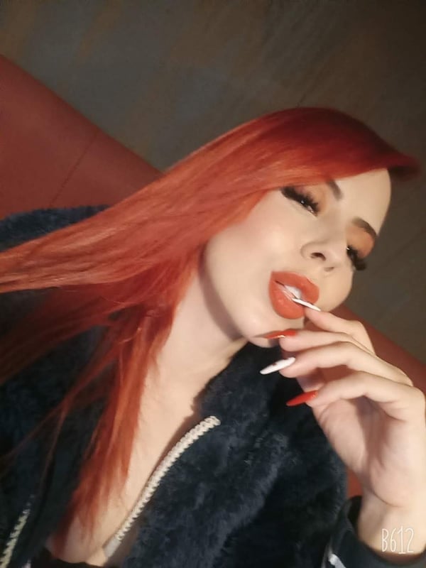 A woman with long red hair posing playfully with a lollipop in her mouth, wearing a black fur jacket against a warm indoor background.