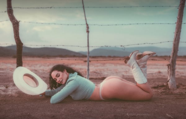 Woman in cowgirl attire with hat and boots posing on the ground near a barbed wire fence in a desert landscape.