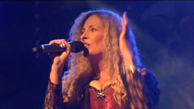 A woman with long curly blonde hair singing passionately into a microphone on stage, wearing a lacy red corset top and dramatic lighting.
