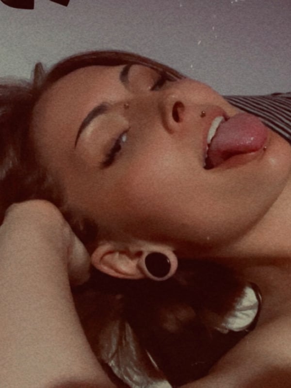 A woman with brown hair and visible ear piercings lying down with her tongue extended, overlaid with heart shapes.