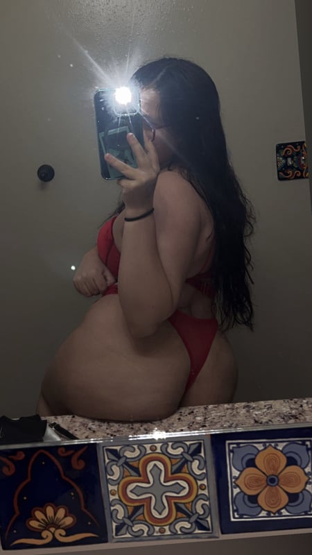 Young woman with long dark hair posing in a red swimsuit in front of a bathroom mirror with colorful tiled accents.