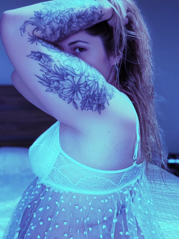 A woman with long blonde hair and floral tattoos on her arms posing in a lace top.