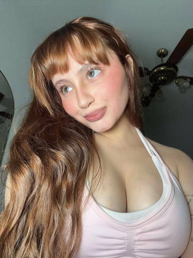 A young woman with long brown hair poses indoors in a light-colored top.
