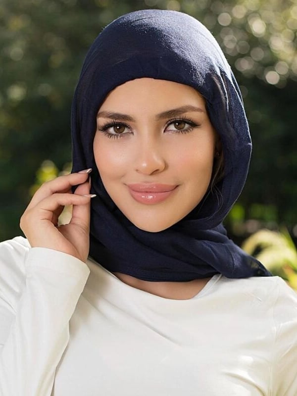 A woman wearing a navy blue headscarf smiles softly outdoors with green foliage blurred in the background.