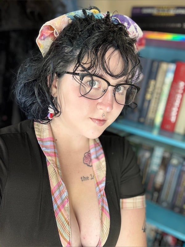 Close-up portrait of a person with curly dark hair, glasses, and colorful headscarf, smiling softly against a bookshelf background.