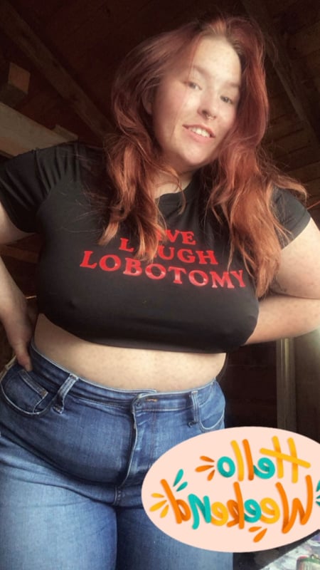A woman with long red hair smiling while wearing a black graphic t-shirt and blue jeans, standing indoors with a colorful sticker nearby.